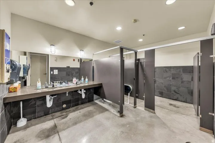 Multi-stall restroom featuring a long vanity with multiple sinks, dark tiled backsplash, and concrete flooring