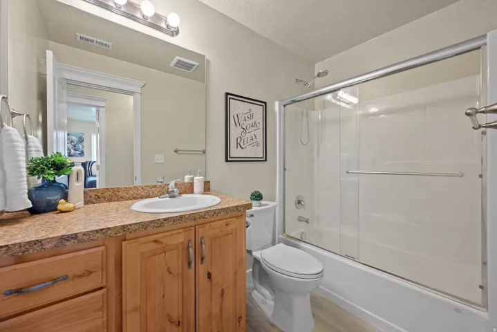 Bathroom featuring a single vanity with wood cabinetry and a speckled countertop, an elongated toilet, and a white tub with a sliding glass door enclosure