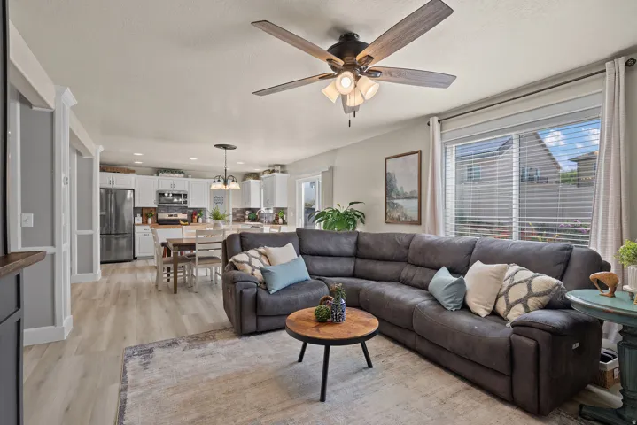 Open concept living area with wood-finish flooring, a ceiling fan with integrated lighting, and a large window with blinds
