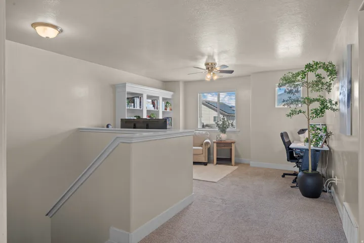 Carpeted upper-level space featuring a built-in bookshelf, ceiling fan, and window with blinds