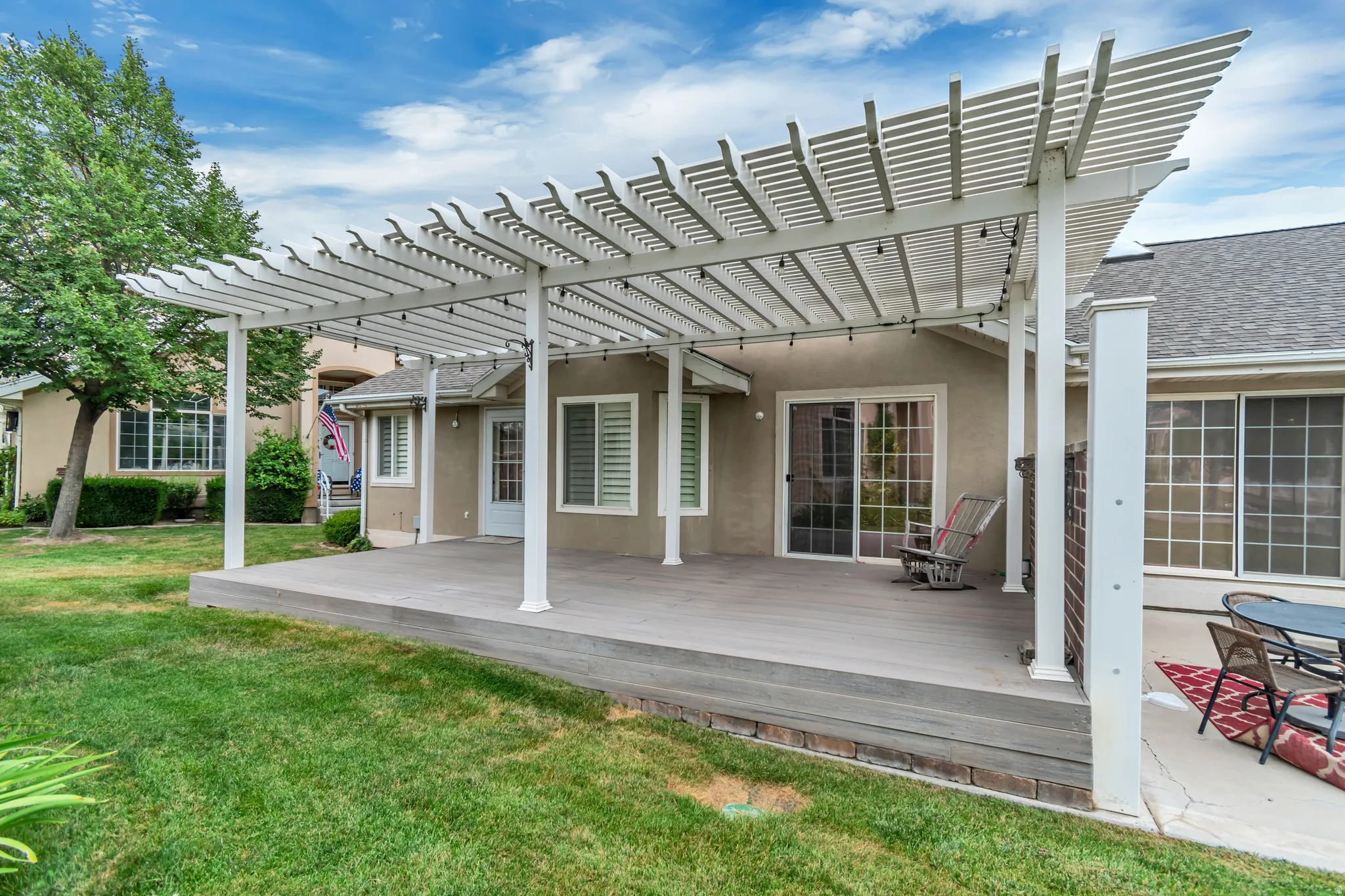 Expansive outdoor living area featuring a large wood-finish deck with a white slatted pergola, string lighting, and sliding glass door access