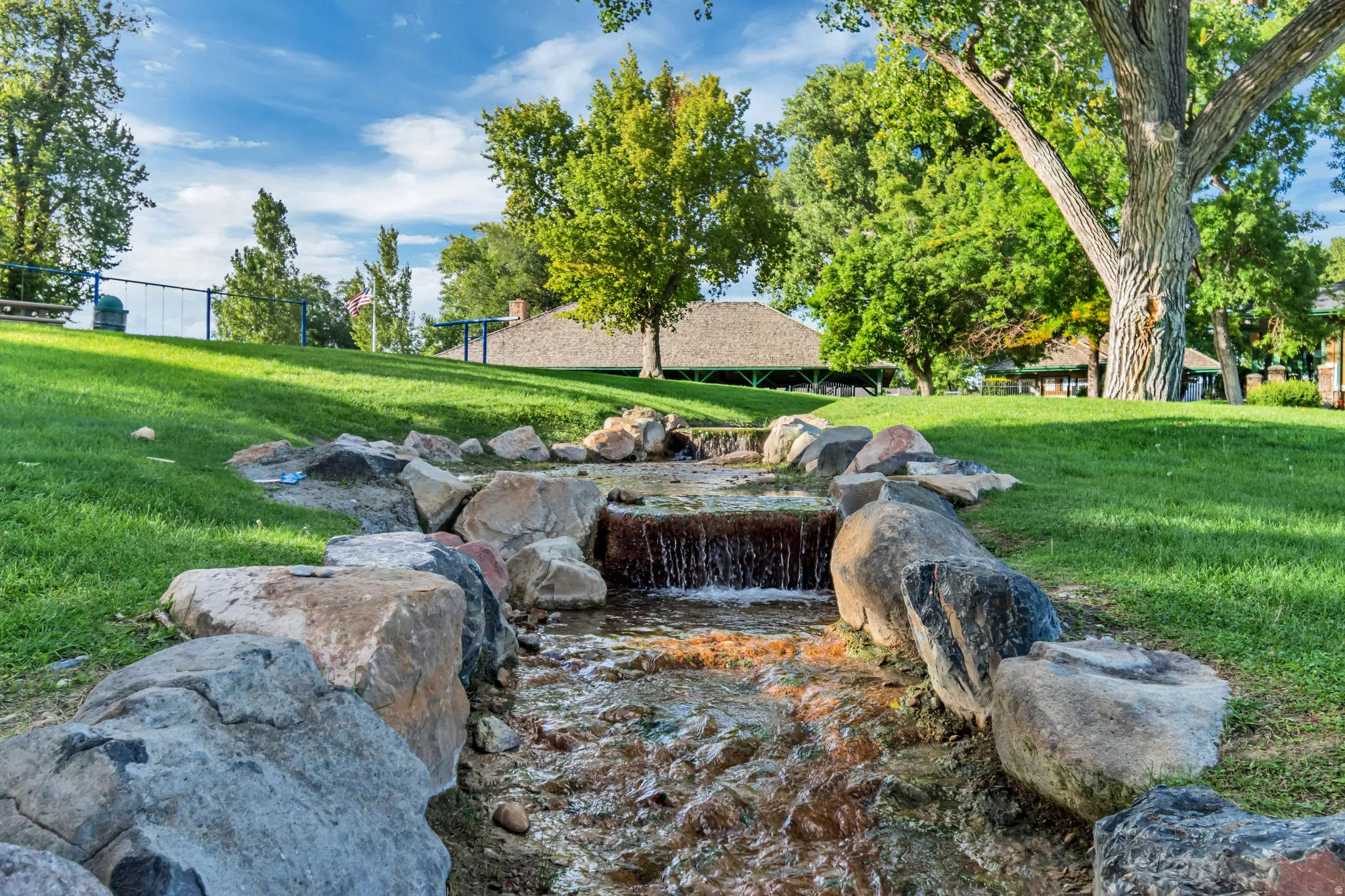 Meandering stream with rock-lined banks, surrounded by a gently sloping lawn and mature trees