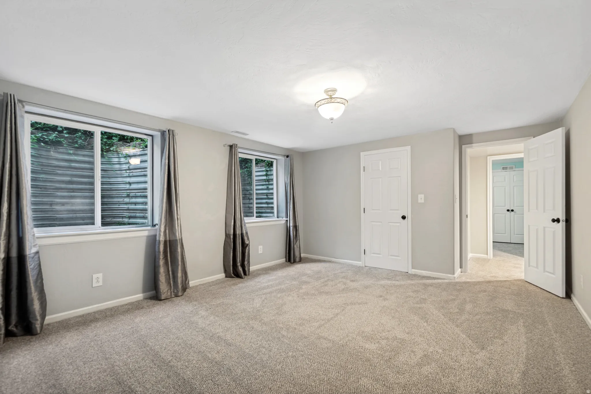 Spacious room featuring light gray walls, plush carpet flooring, two windows, and multiple paneled doors