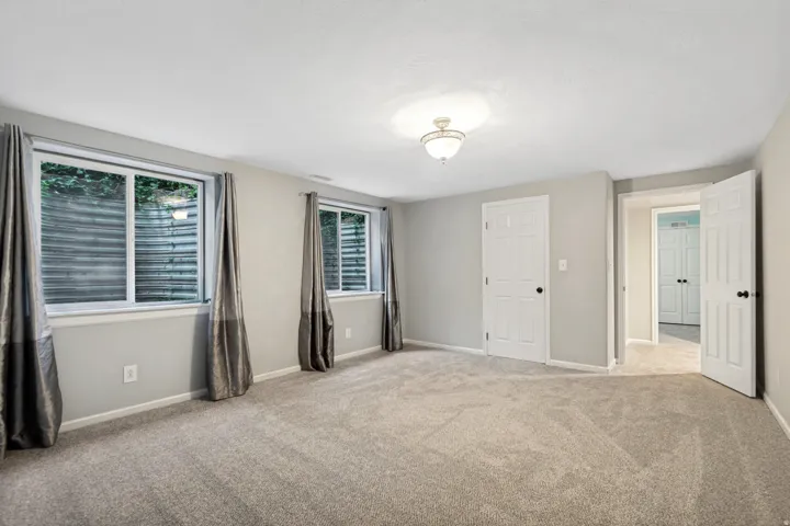 Spacious room featuring light gray walls, plush carpet flooring, two windows, and multiple paneled doors