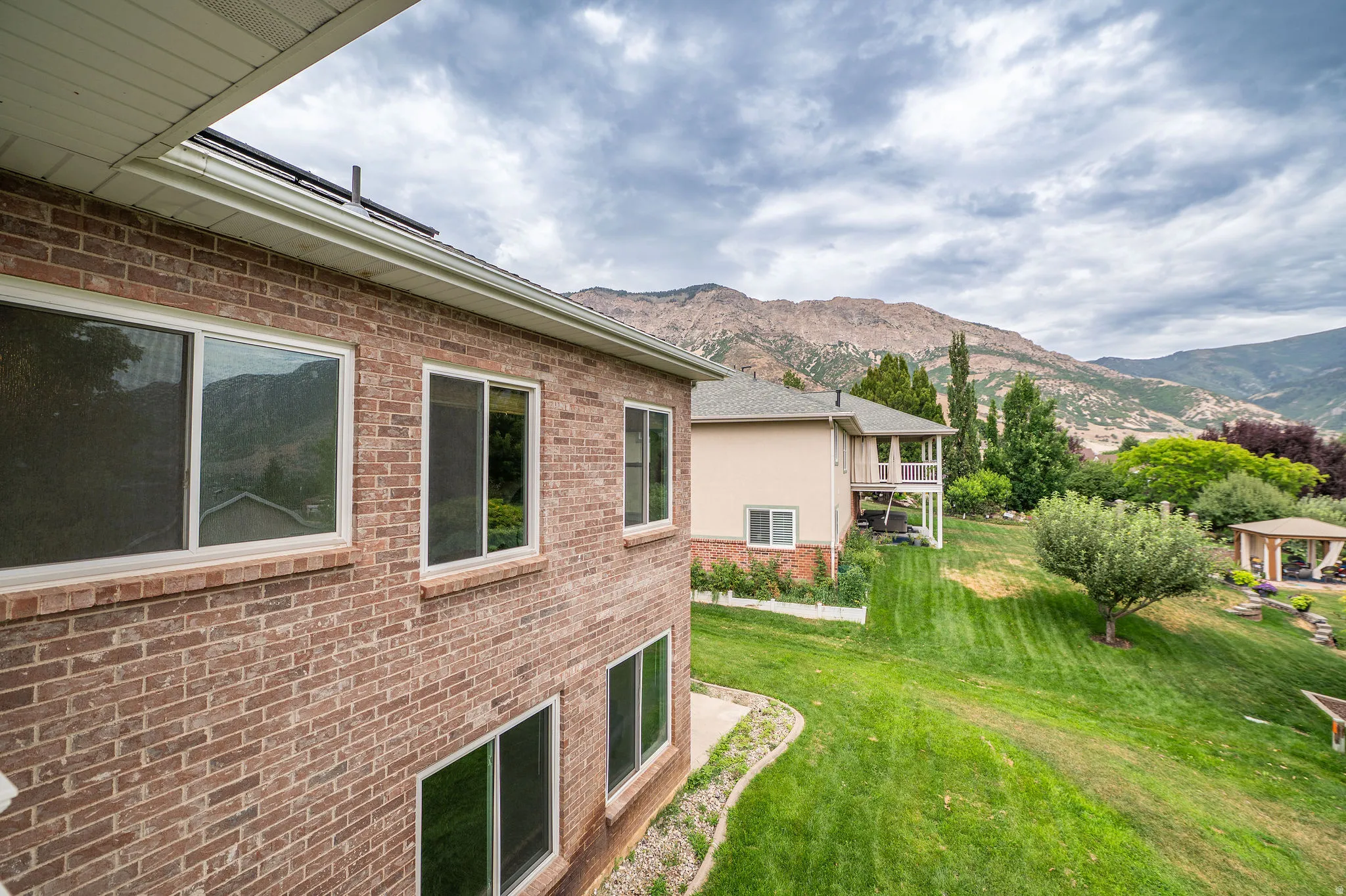 Brick exterior with white-framed windows, a sloping green lawn, a covered patio structure, and distant mountains