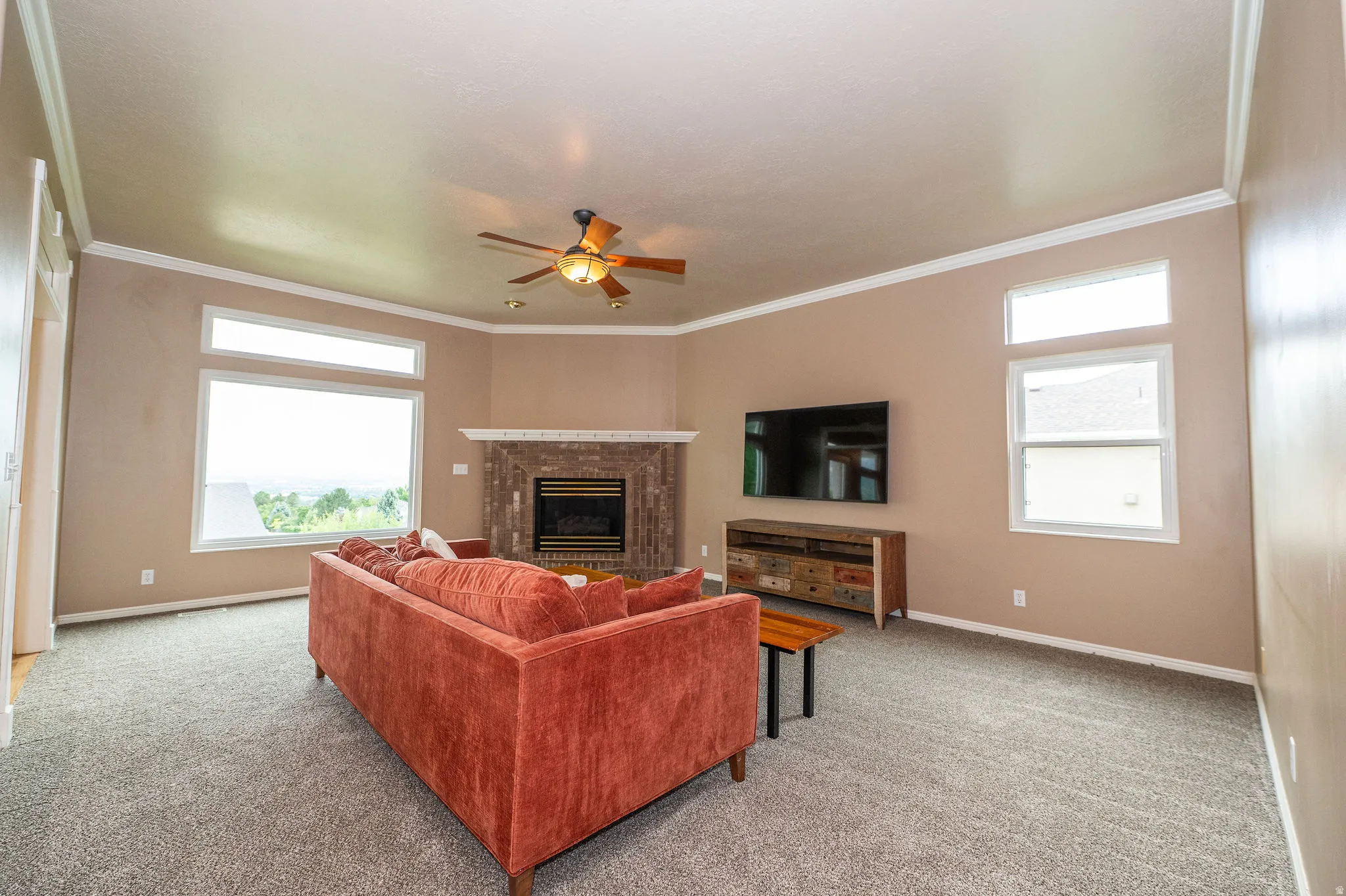 Living area featuring a corner brick fireplace, multiple windows, a ceiling fan with integrated lighting, crown molding, and carpet flooring