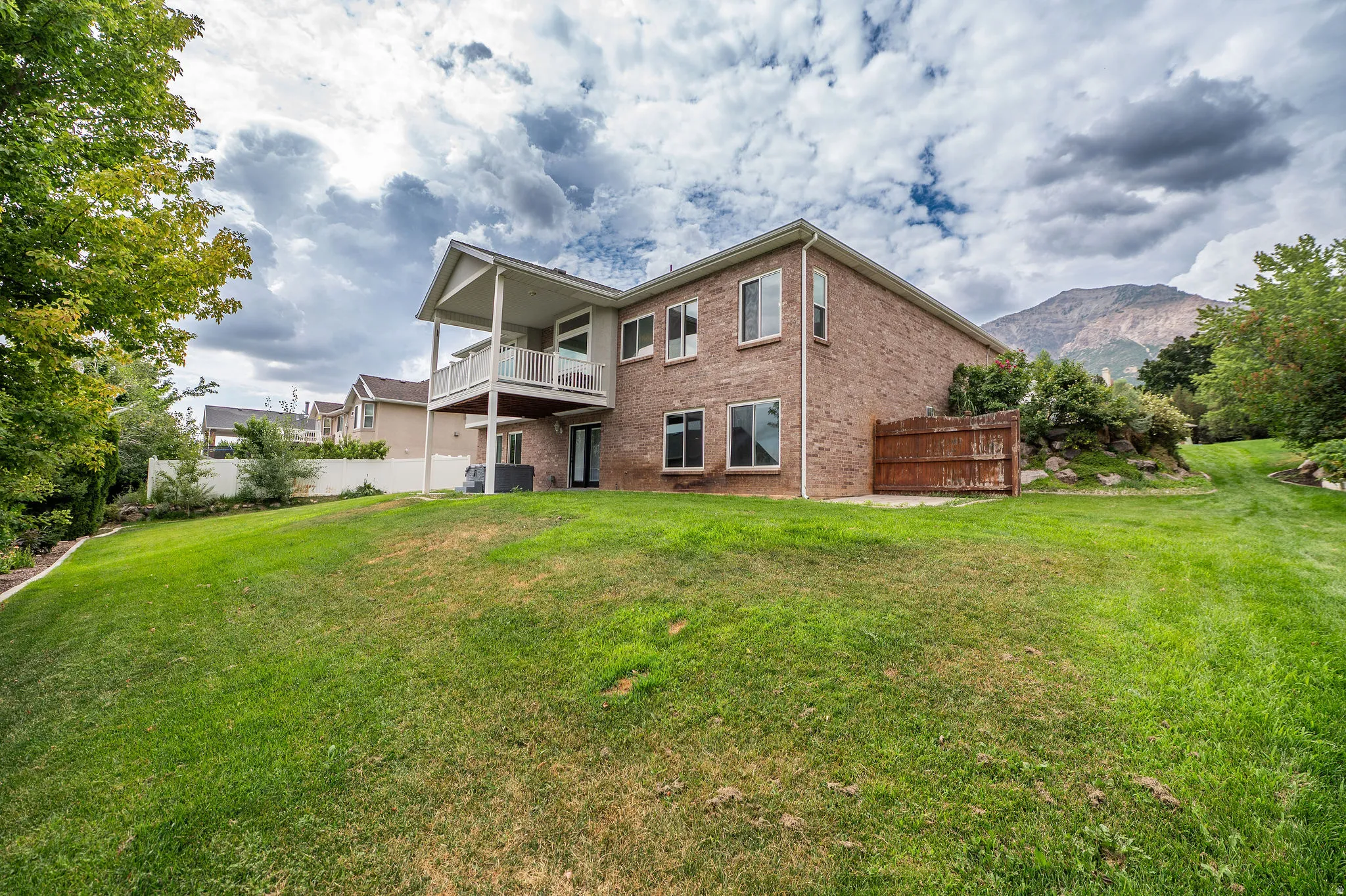 Expansive sloped lawn area featuring a two-story brick exterior, upper-level covered balcony, lower-level patio access, and mature trees