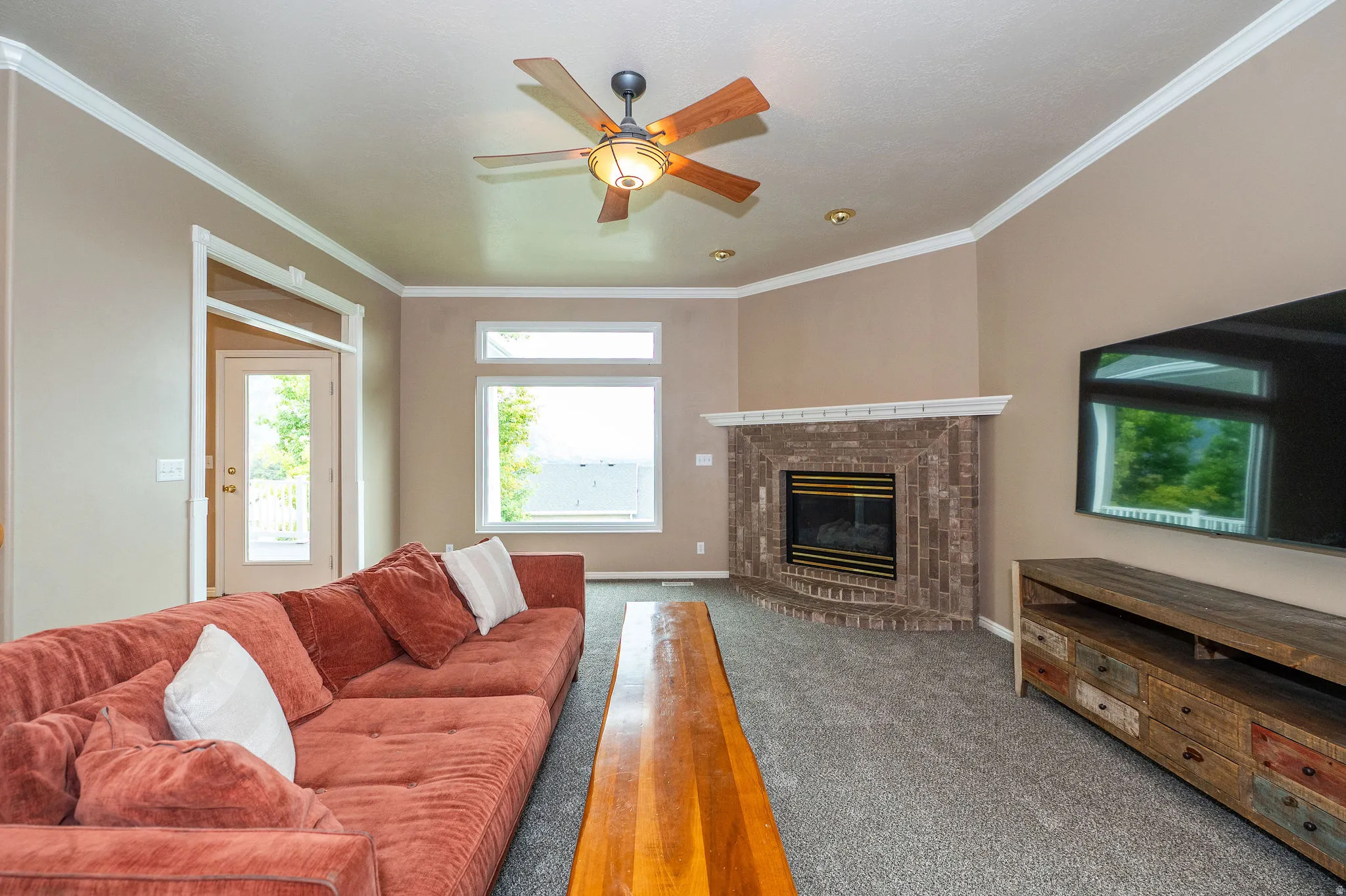 Spacious living area featuring carpet flooring, a corner brick fireplace with a white mantel, and a ceiling fan with integrated lighting