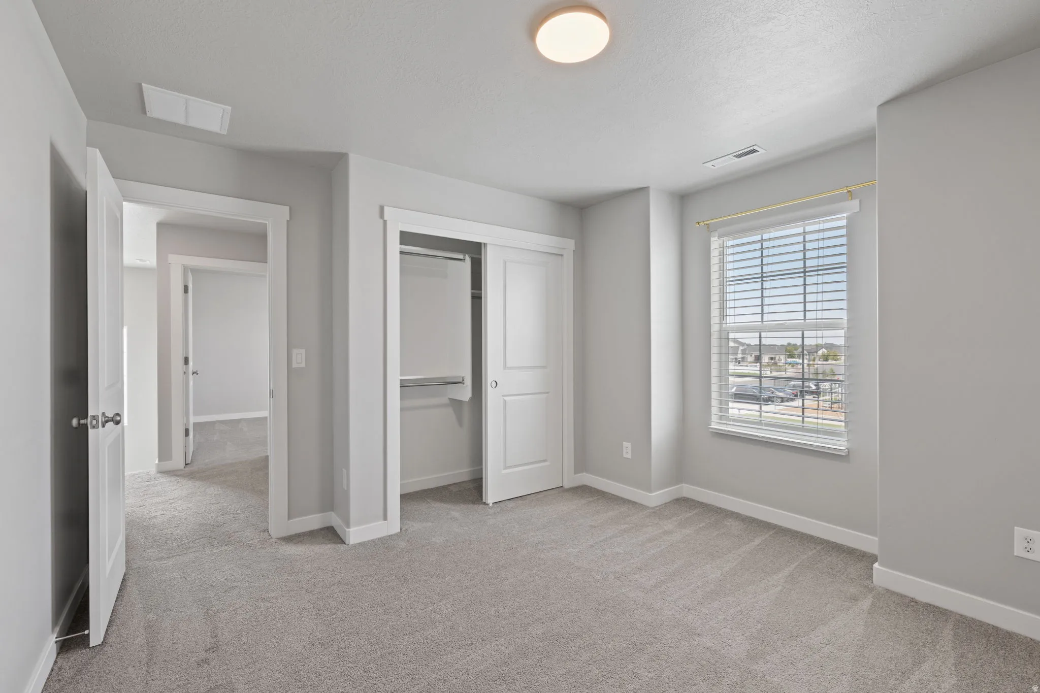 Spacious room featuring light gray carpet, neutral wall paint, a double-door closet with shelving, a window with horizontal blinds, and overhead flush mount lighting