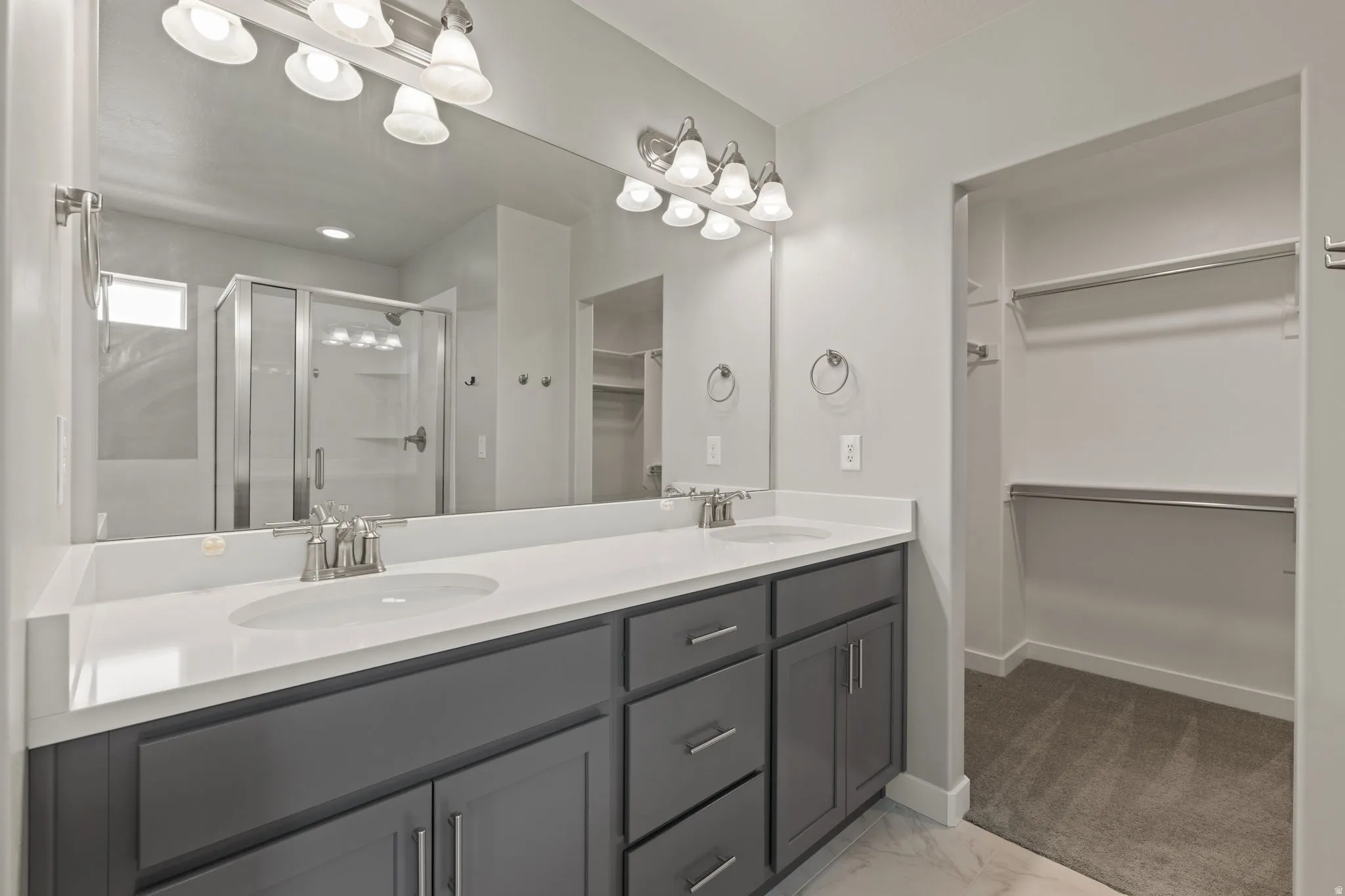 Bathroom featuring a dual vanity with a white countertop and gray cabinetry, a large wall-mounted mirror, a glass-enclosed shower, and a walk-in closet with built-in shelving and hanging rods
