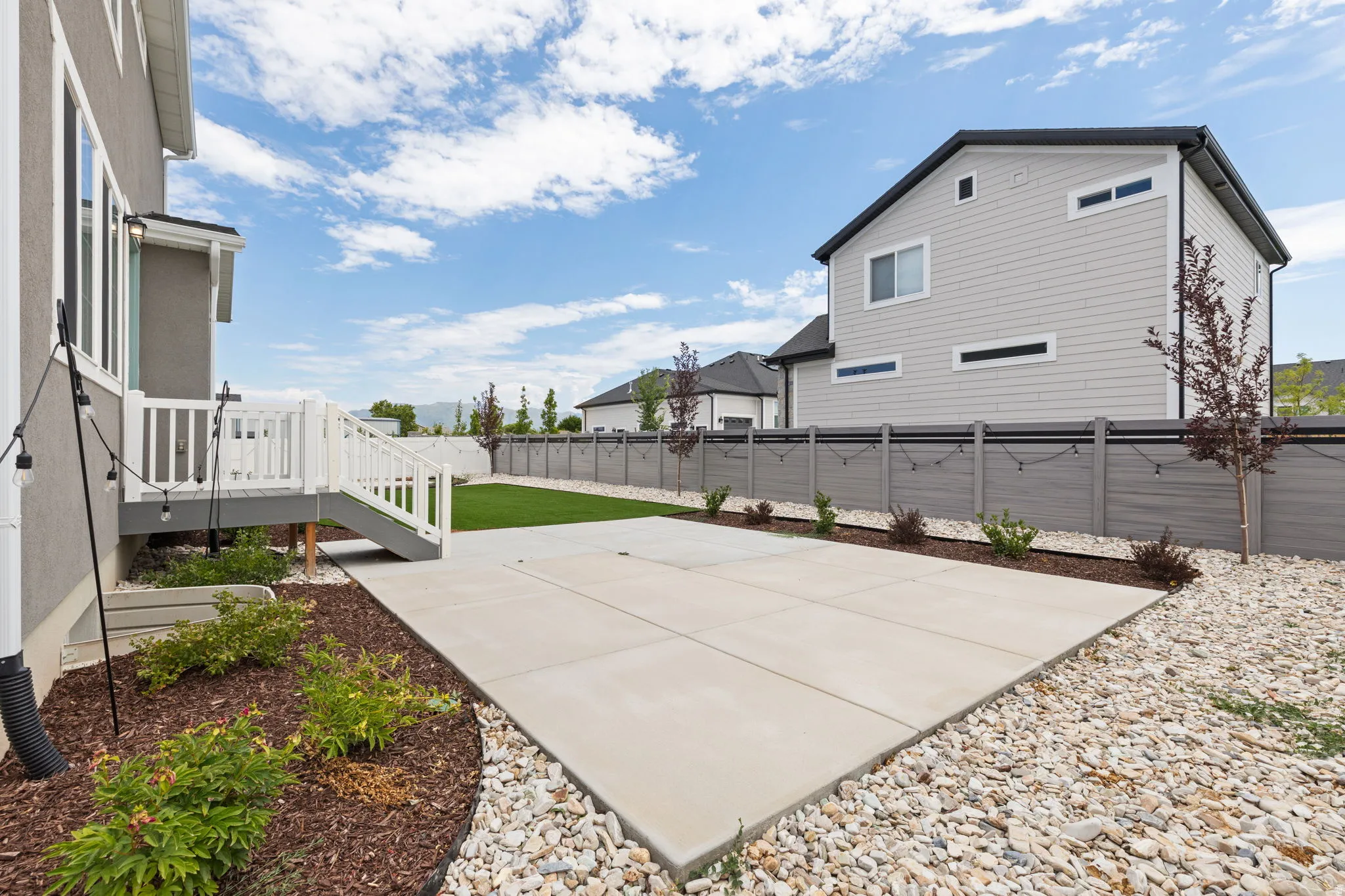 Expansive backyard featuring a concrete patio, landscaped beds with mulch and plantings, and a white deck with stairs