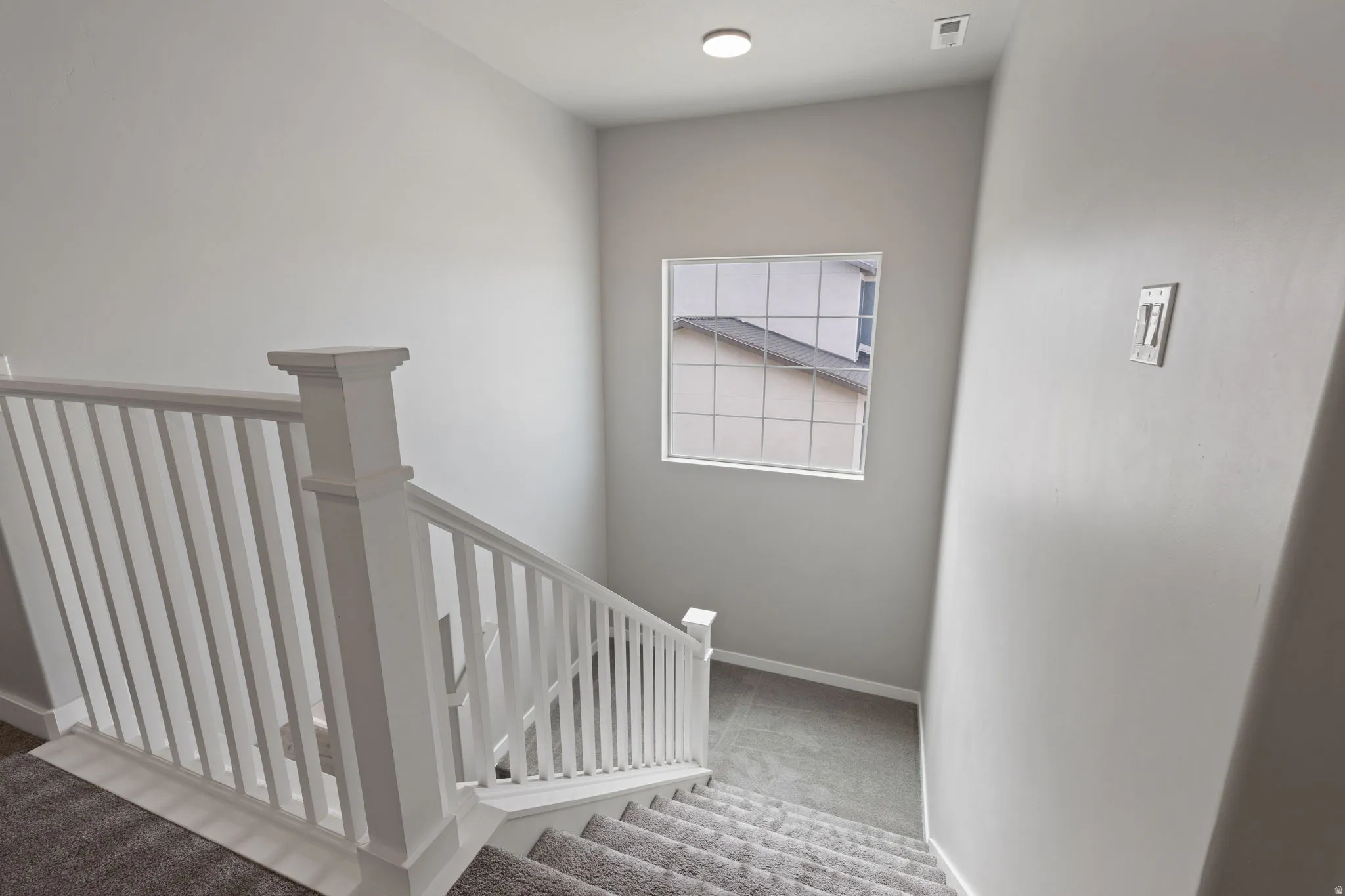 Staircase featuring white balusters and newel posts, gray carpeted treads, and a square window with grid detailing