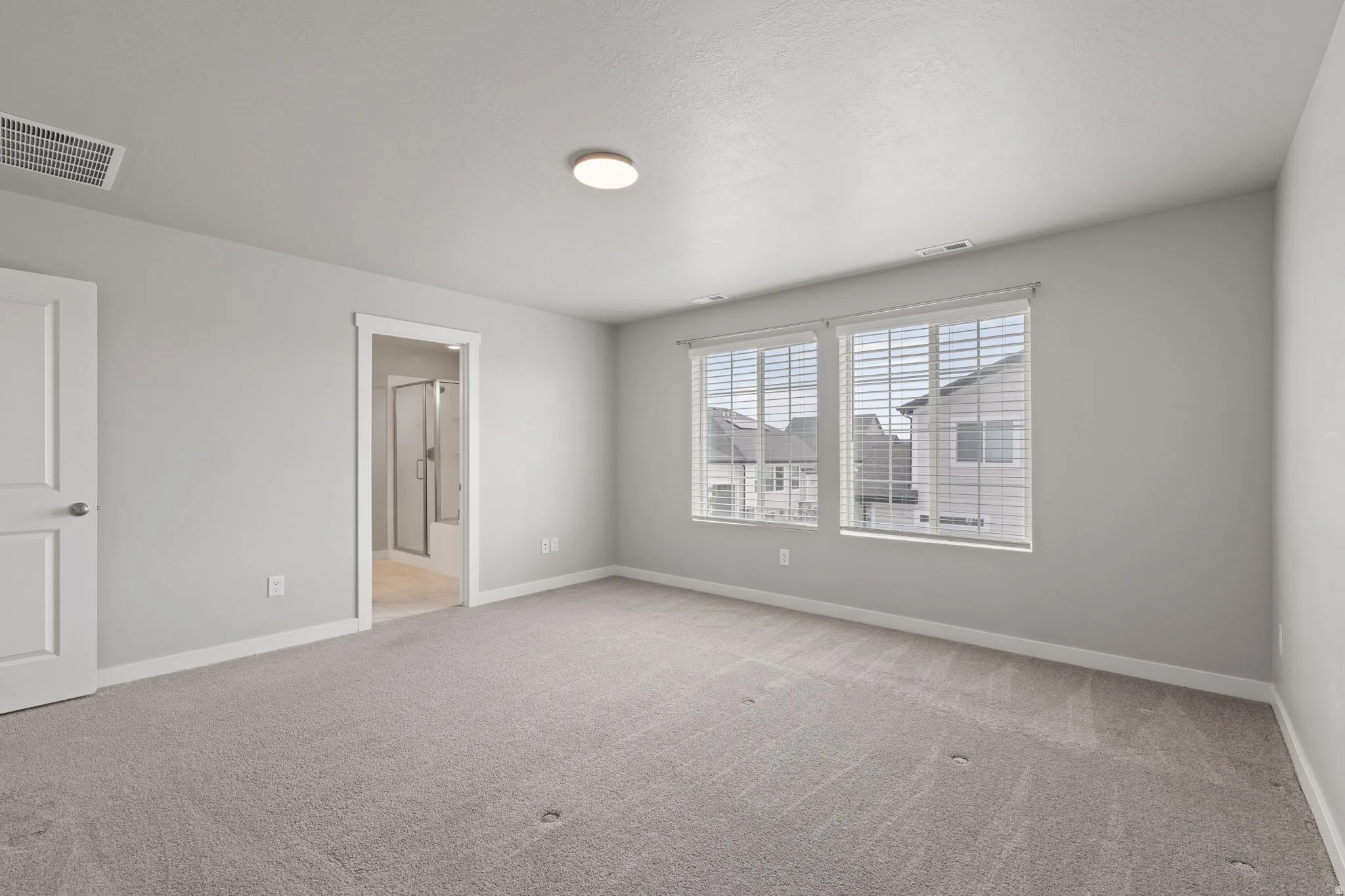 Carpeted room with a ceiling-mounted light fixture, dual windows with blinds, a white paneled door, and an entryway featuring a bathroom with a shower