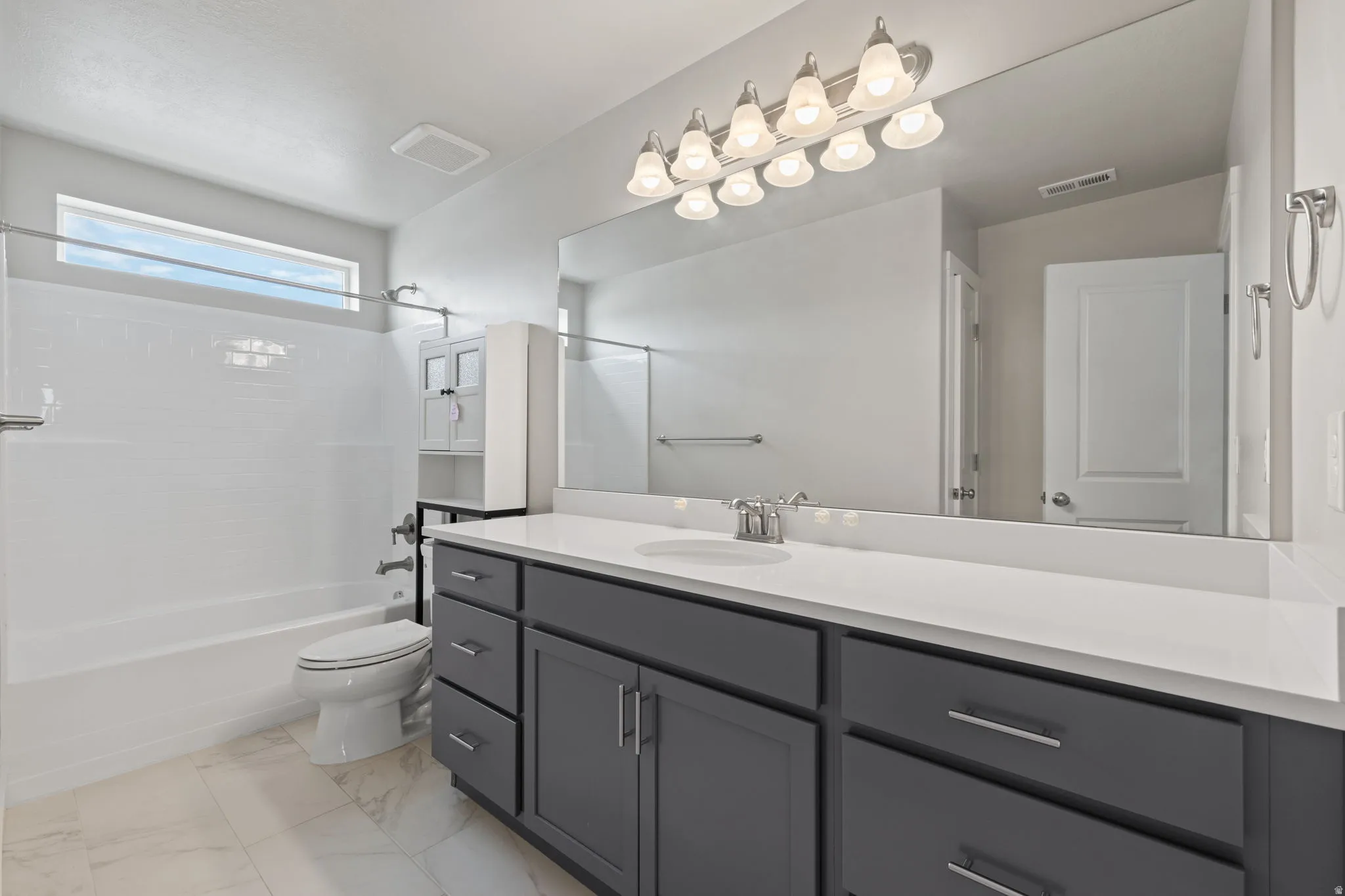 Contemporary bathroom featuring a dark gray double vanity with white countertops, expansive mirror, a combination tub and shower with white subway tile surround, and white tile flooring