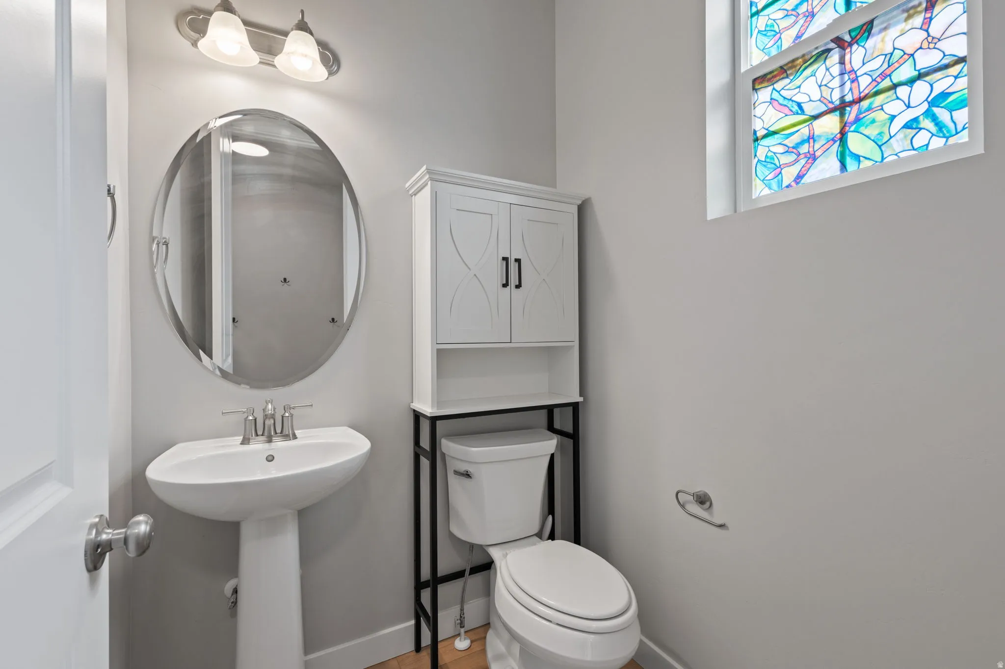 Pedestal sink with a brushed nickel faucet, oval mirror, and a stained-glass window