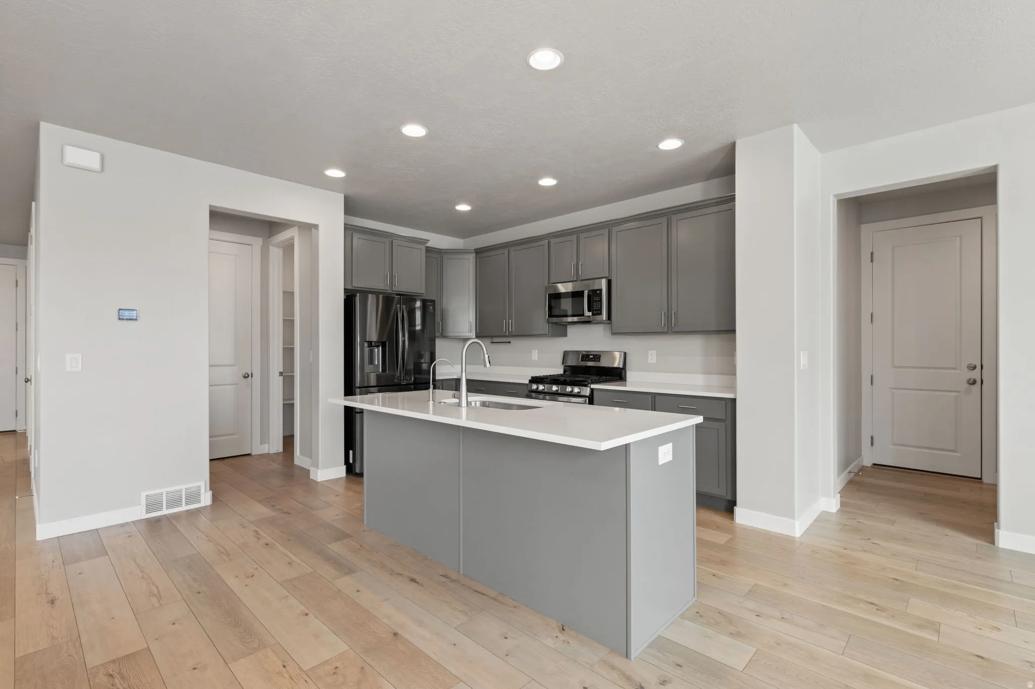 Modern kitchen featuring a central island with a sink, white countertops, gray cabinetry, and stainless steel appliances