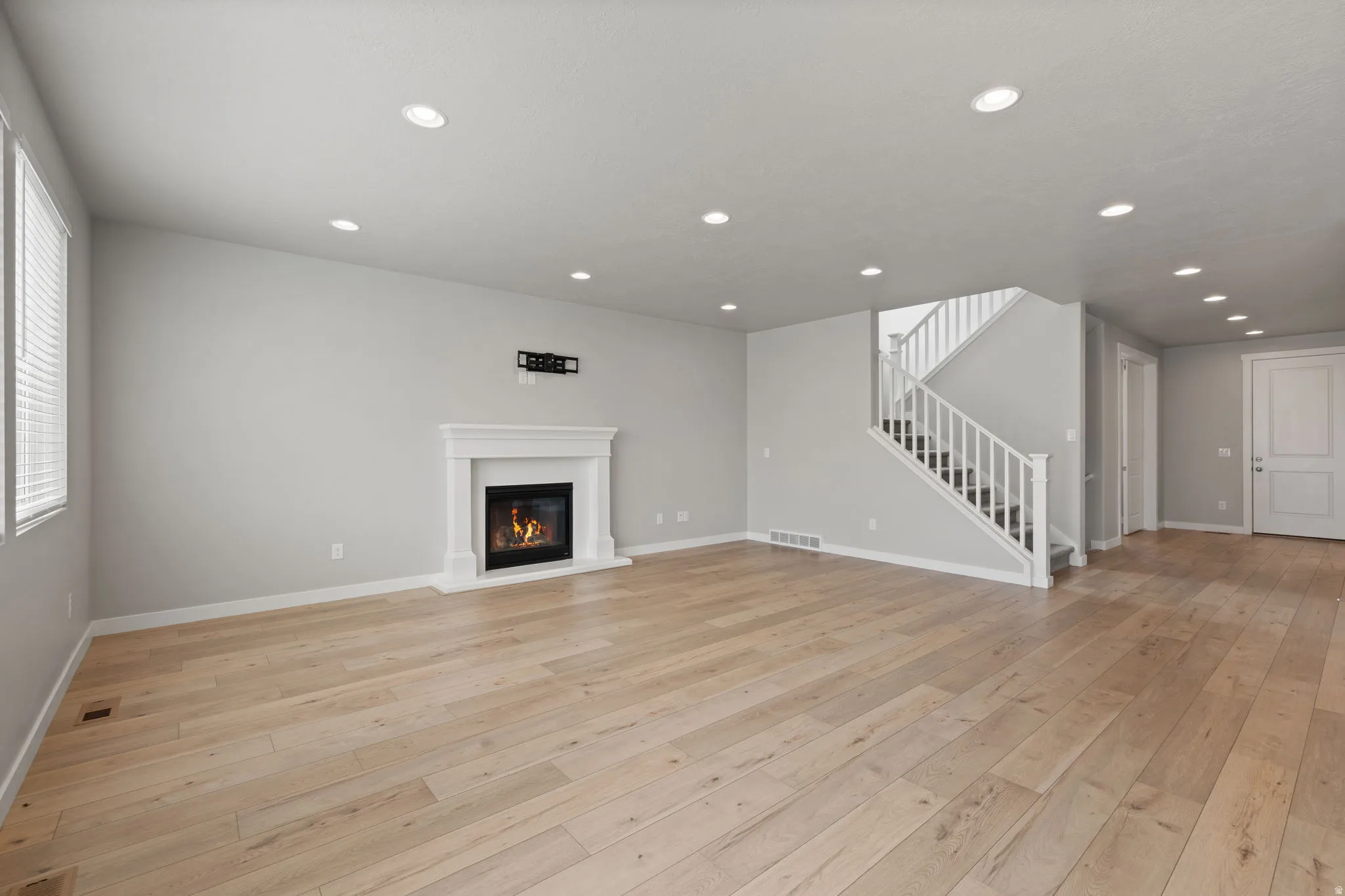 Spacious living area featuring wood-finish flooring, a contemporary fireplace with a white mantel, recessed ceiling lighting, and a prominent staircase with white railings