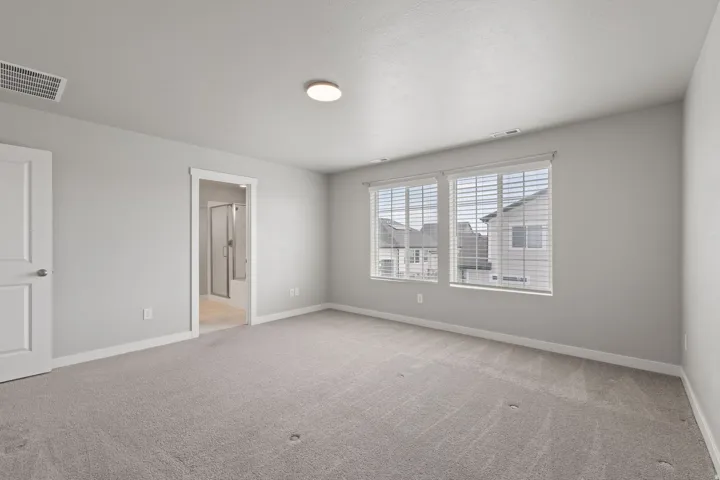 Carpeted room with a ceiling-mounted light fixture, dual windows with blinds, a white paneled door, and an entryway featuring a bathroom with a shower