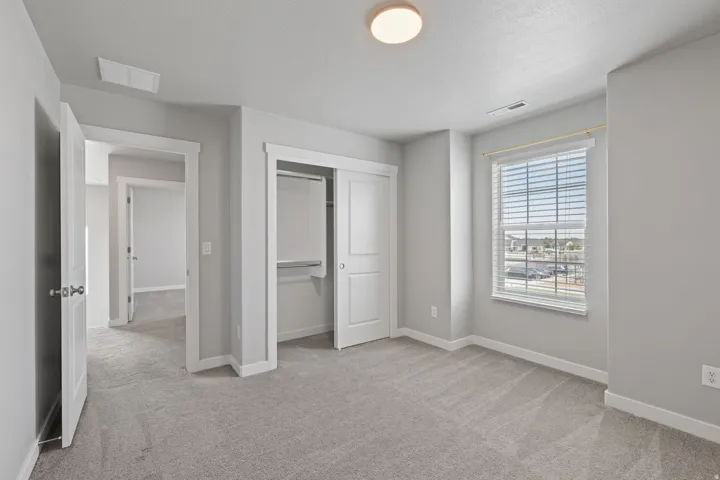 Spacious room featuring light gray carpet, neutral wall paint, a double-door closet with shelving, a window with horizontal blinds, and overhead flush mount lighting