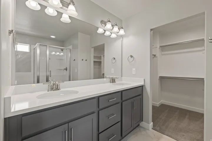 Bathroom featuring a dual vanity with a white countertop and gray cabinetry, a large wall-mounted mirror, a glass-enclosed shower, and a walk-in closet with built-in shelving and hanging rods