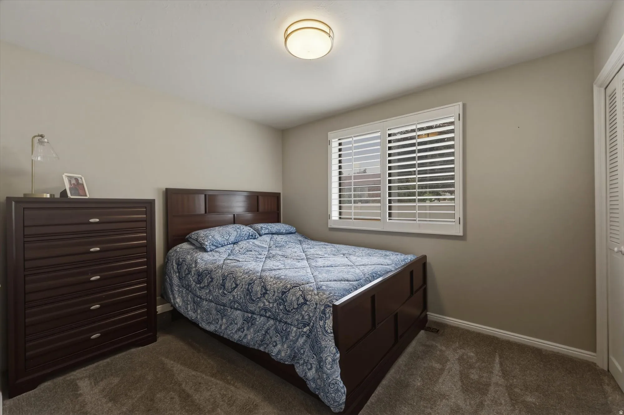 Carpeted room featuring neutral-toned walls, a window with plantation shutters, a ceiling-mounted light fixture, and a louvered bi-fold door