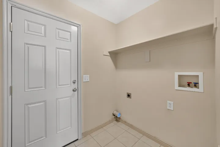 Utility room featuring tile flooring, a white paneled door, a wall-mounted shelf, and laundry connections