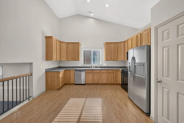 Vaulted ceiling kitchen featuring wood-finish flooring, ample wood cabinetry, a stainless steel refrigerator, and a built-in dishwasher