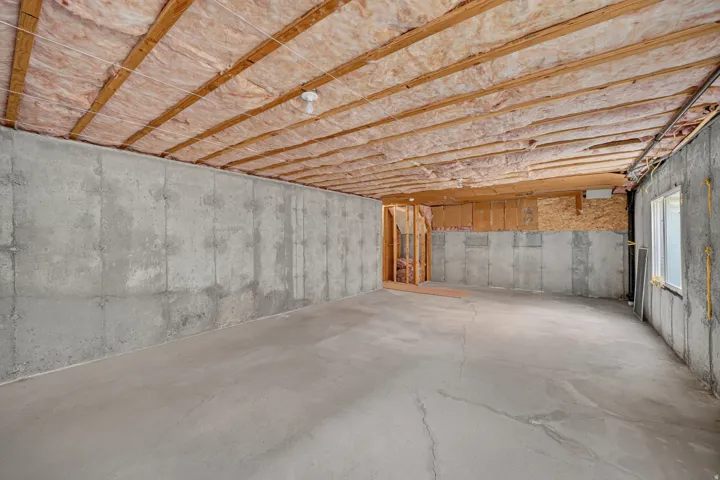 Expansive unfinished basement featuring concrete walls and flooring, exposed ceiling joists with insulation, and a window for natural light