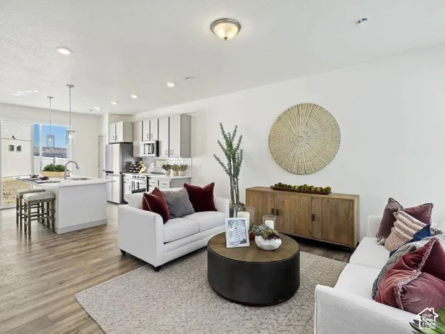 Open-concept living space featuring wood-finish flooring, recessed lighting, white walls, a kitchen island with a sink, and stainless steel appliances