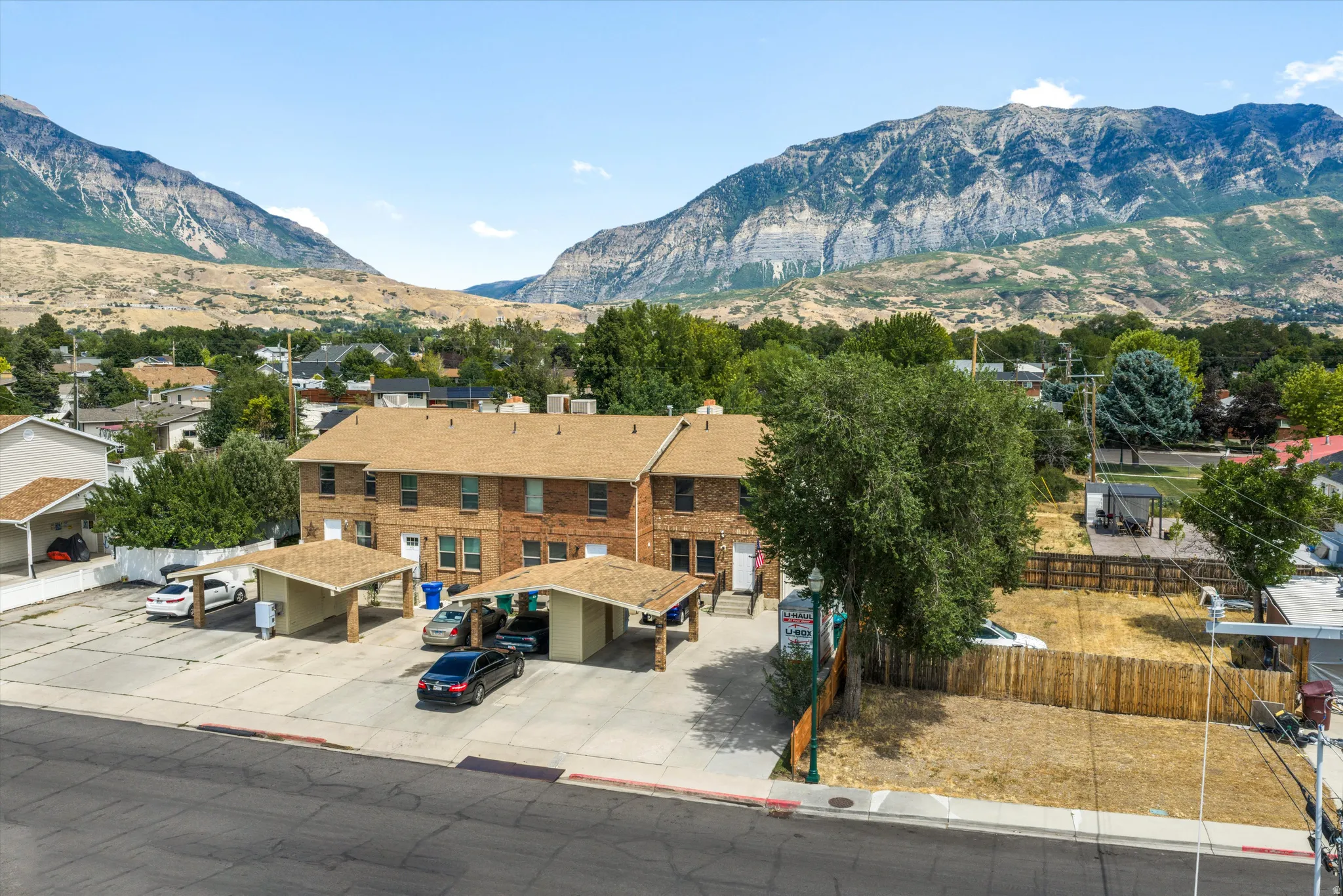 Brick multi-unit property featuring a multi-car parking lot with two covered carports, individual unit entrances, and a partial wood fence