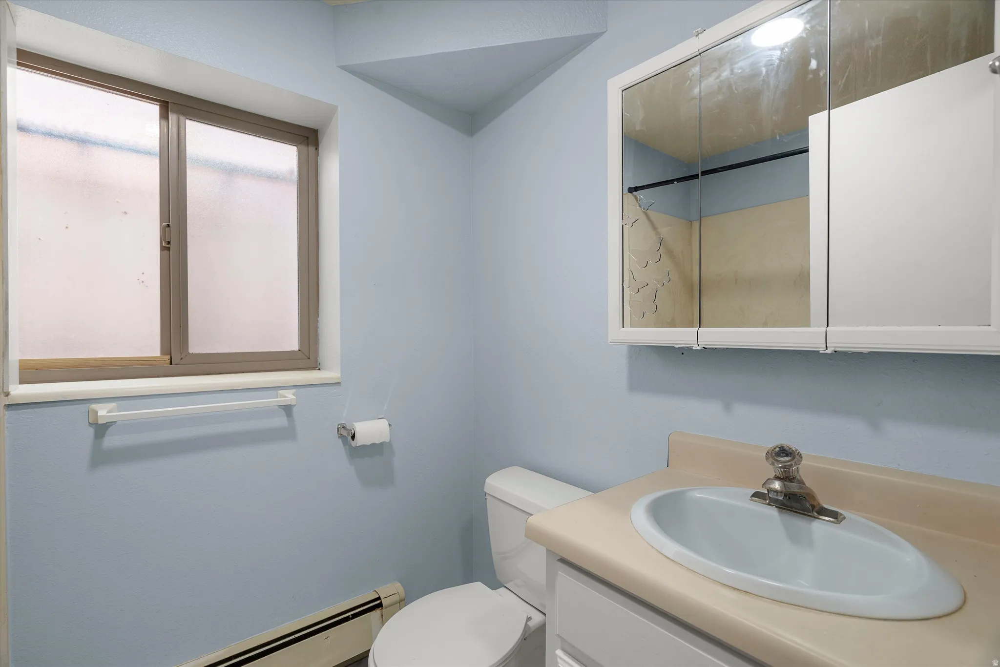 Bathroom featuring a sliding window with frosted glass, a wall-mounted medicine cabinet with mirrored doors, a single vanity with an integrated sink, and a toilet