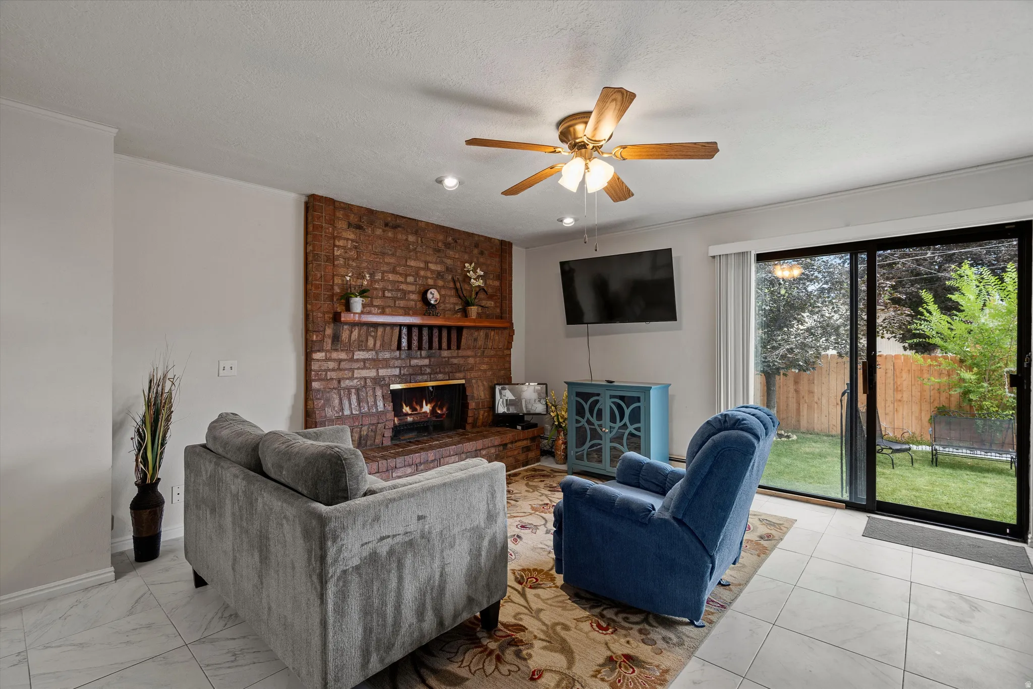 Brick fireplace with wood mantel, ceiling fan, sliding glass door, and tile flooring