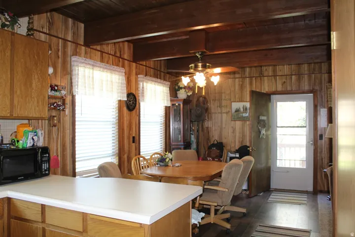Open concept kitchen and dining area with wood-finish paneling, exposed wood beams, ceiling fan with light fixture, and two bright windows