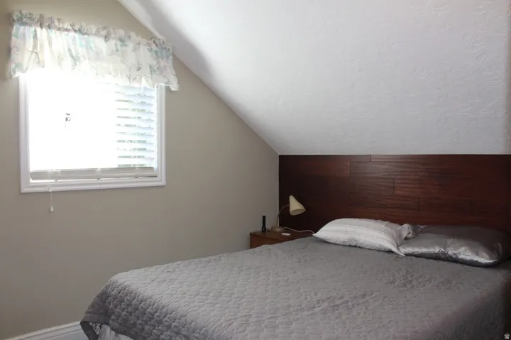 Sloped ceiling room featuring a window with blinds, a dark wood-finish headboard, and light neutral walls