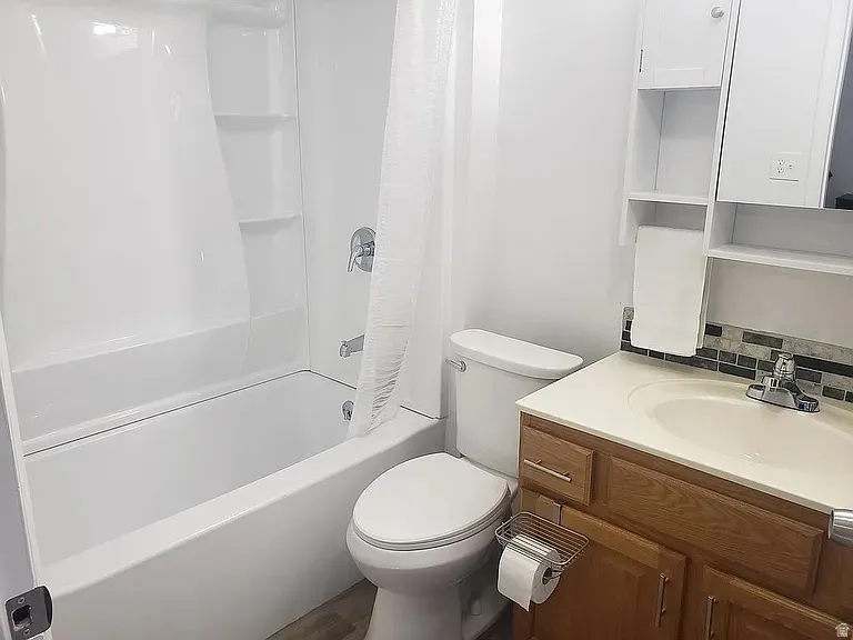 Bathroom featuring a white bathtub and shower surround with built-in shelving, a toilet, a wooden vanity with a light-colored countertop and integrated sink, a mosaic tile backsplash, and an overhead medicine cabinet with open shelving