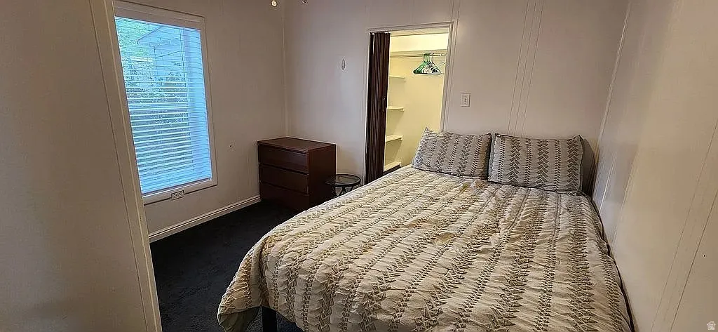Bedroom featuring a window with horizontal blinds, dark carpeting, and a built-in closet with shelving and bi-fold doors