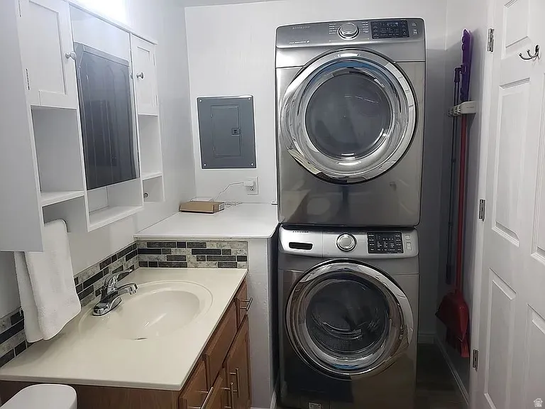 Laundry area featuring a stacked washer and dryer, an integrated sink, a mosaic tile backsplash, white upper cabinetry with open shelving, and a wood-finish vanity base