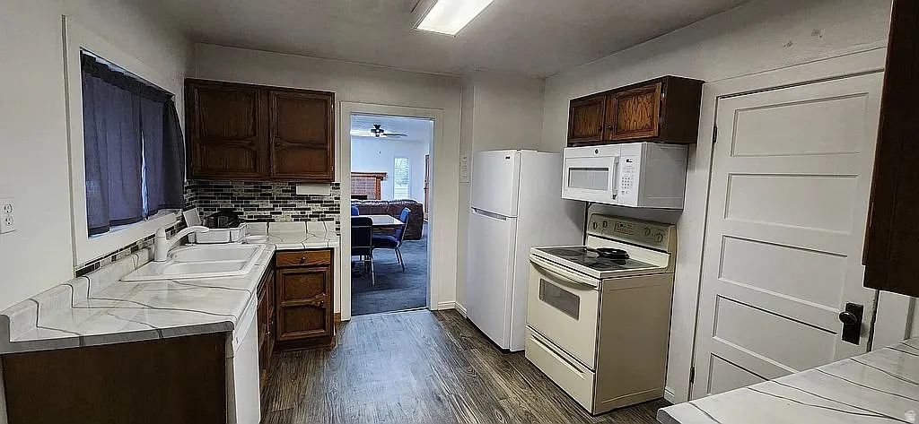 Kitchen featuring wood-finish flooring, white countertops with gray veining, dark wood cabinetry, a mosaic tile backsplash, and white appliances
