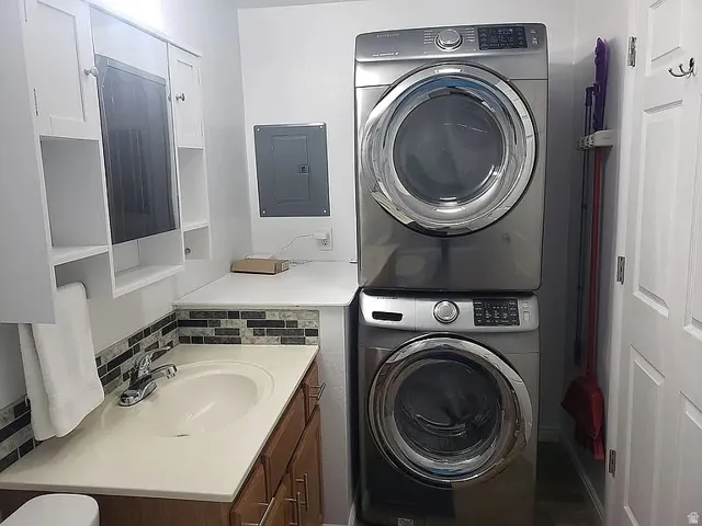 Laundry area featuring a stacked washer and dryer, an integrated sink, a mosaic tile backsplash, white upper cabinetry with open shelving, and a wood-finish vanity base