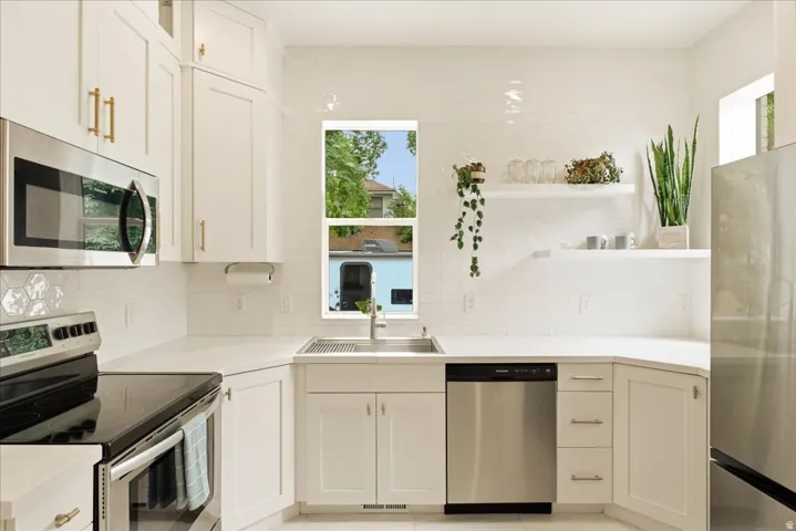 Modern kitchen featuring white cabinetry with gold-tone hardware, stainless steel appliances, white countertops, and a full-height tile backsplash