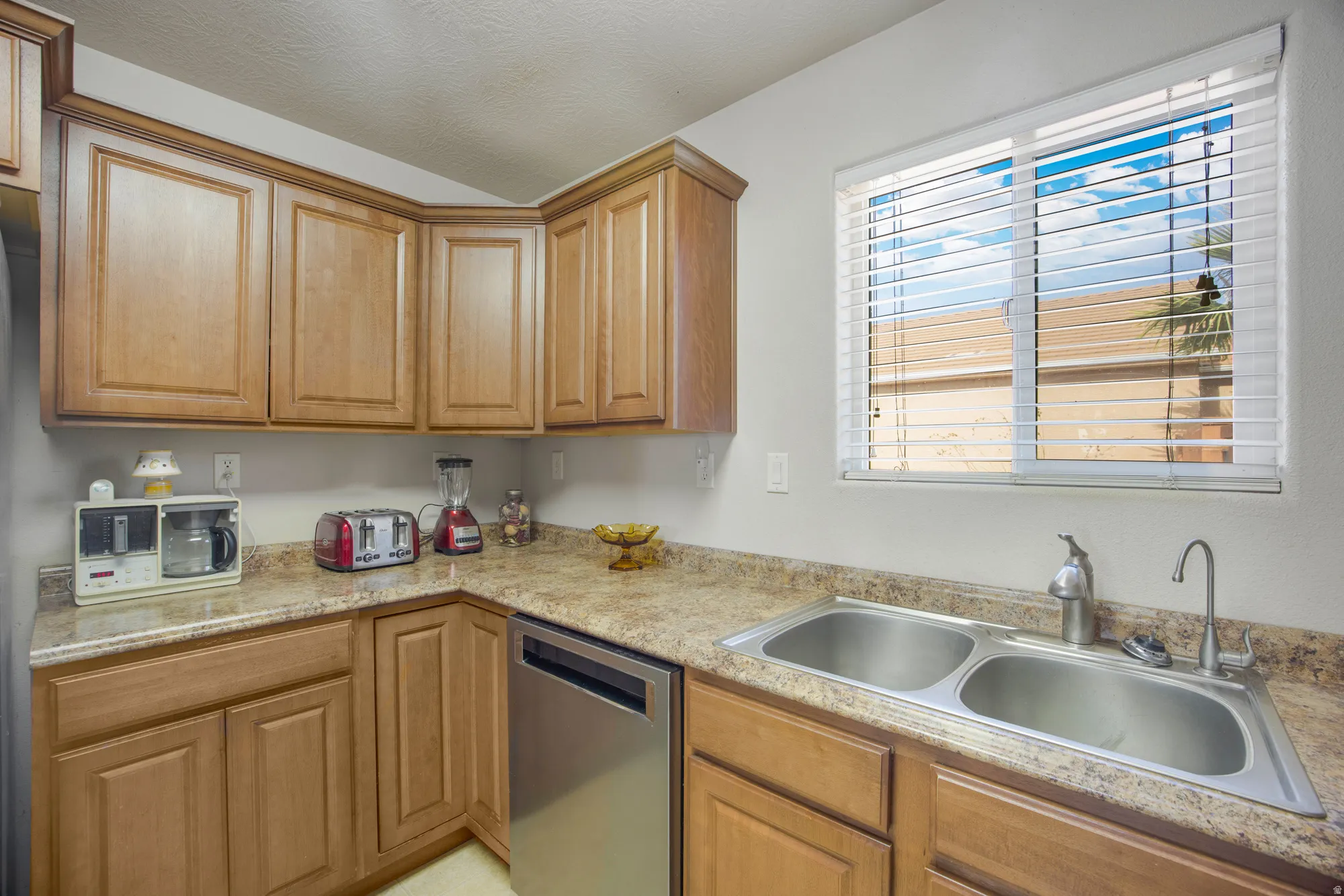 Kitchen featuring light wood cabinetry, a double basin stainless steel sink, a stainless steel dishwasher, a light-toned patterned countertop, and a window with horizontal blinds