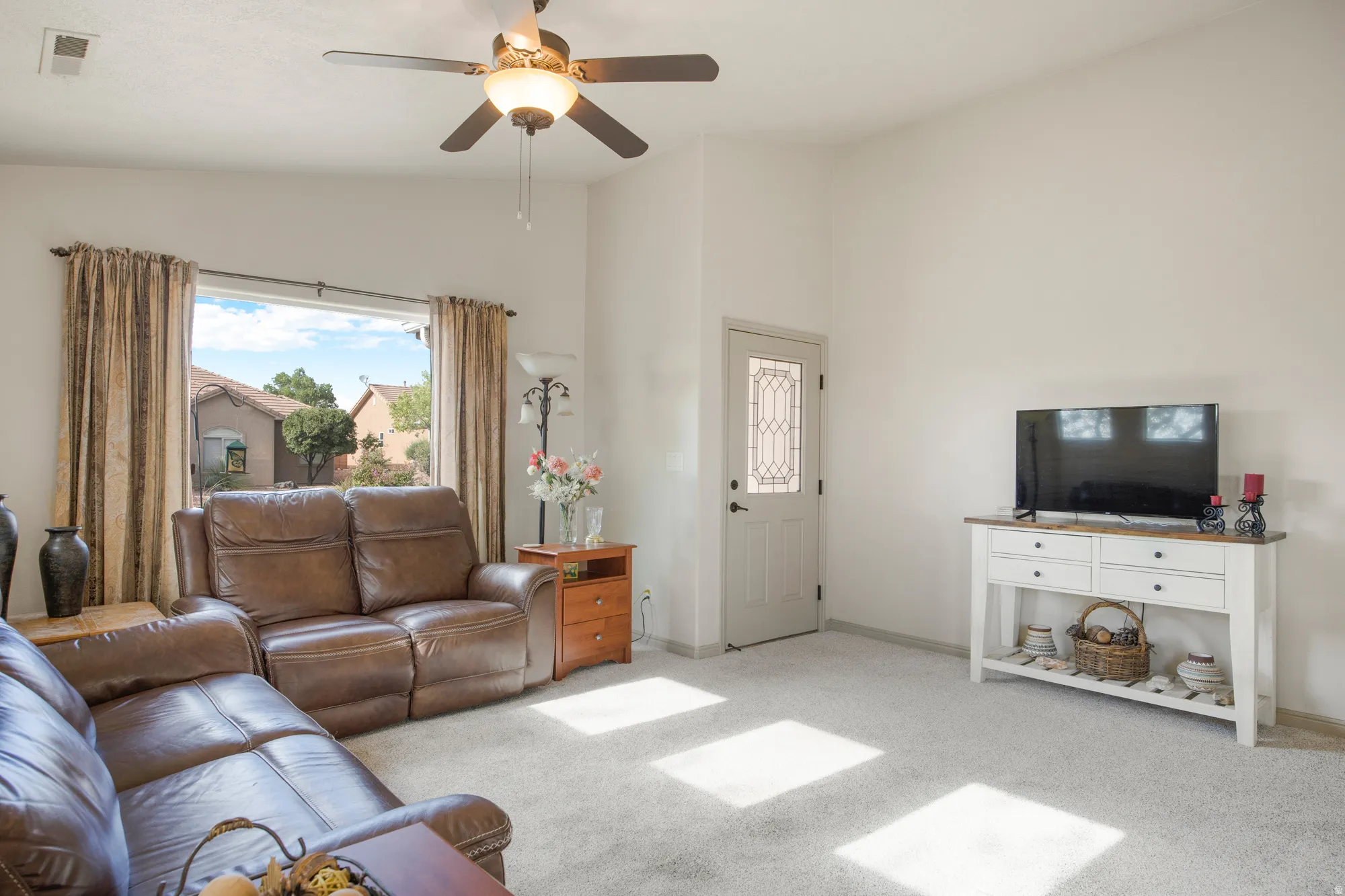 Spacious living area featuring light gray carpeting, tall white walls, and a ceiling fan with integrated lighting