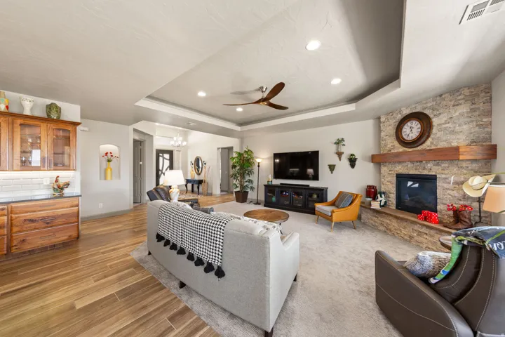 Spacious living area featuring a stacked stone fireplace with a substantial wood mantel, a tray ceiling with recessed lighting and a ceiling fan, and light wood-finish flooring extending into the kitchen area
