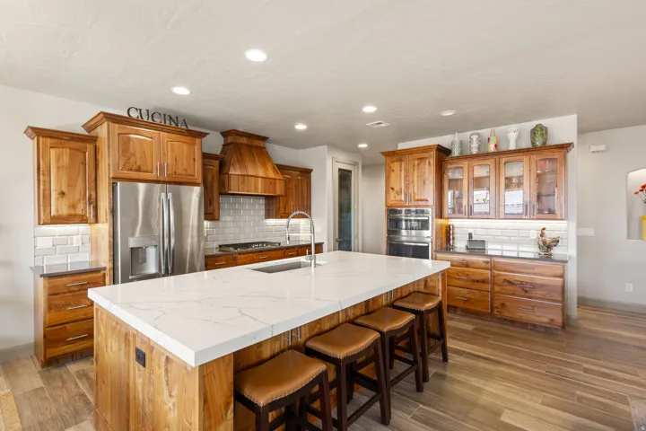 Spacious kitchen featuring a large central island with a white stone countertop, extensive wood cabinetry, stainless steel appliances, a tile backsplash, and wood-finish flooring