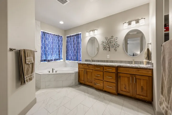 Spacious bathroom featuring a corner soaking tub, dual vanities with granite countertops, wood-finish cabinetry, and contemporary light fixtures