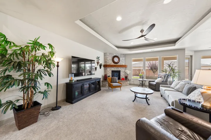 Spacious living area featuring a stone-clad fireplace with wood mantel, a multi-tiered tray ceiling with recessed lighting and a ceiling fan, and large windows