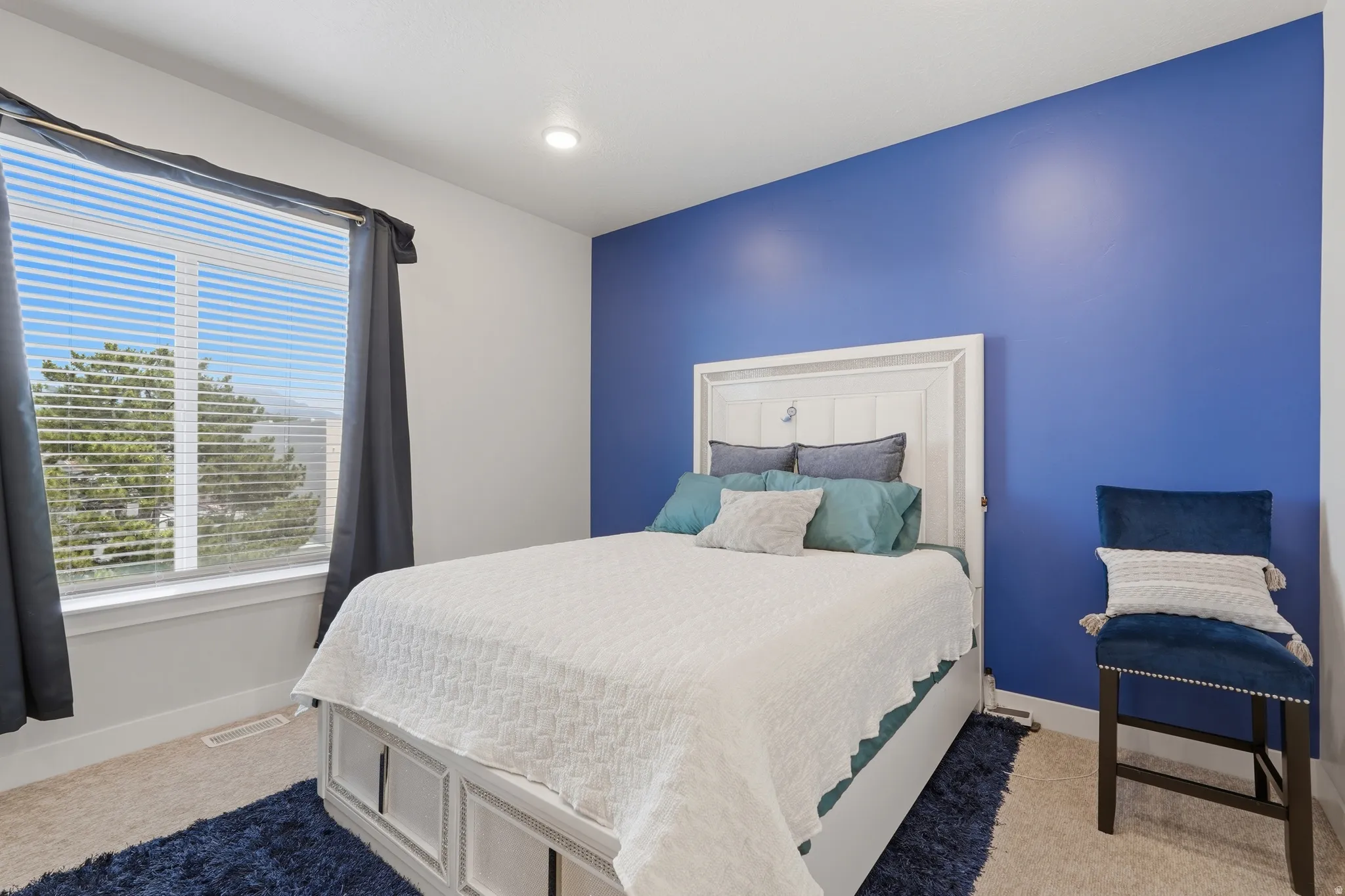 Carpeted bedroom featuring a large window with horizontal blinds, a vibrant blue accent wall, and a white paneled headboard