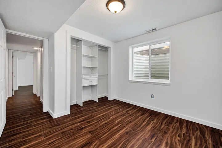 Room featuring wood-finish flooring, a built-in closet system with shelving and drawers, white trim, and a window providing natural light