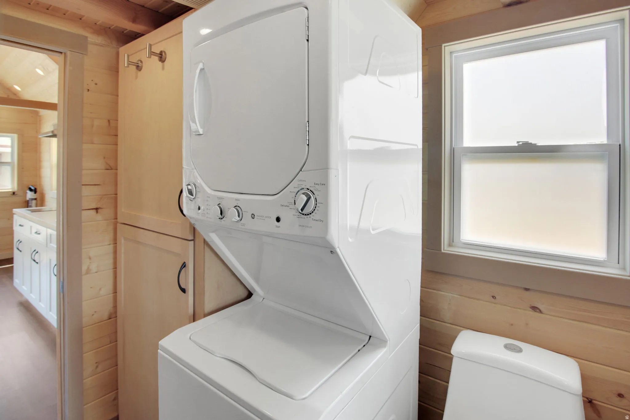 Stackable washer and dryer unit, wood-paneled walls, built-in storage cabinet, white single-hung window, and a white toilet