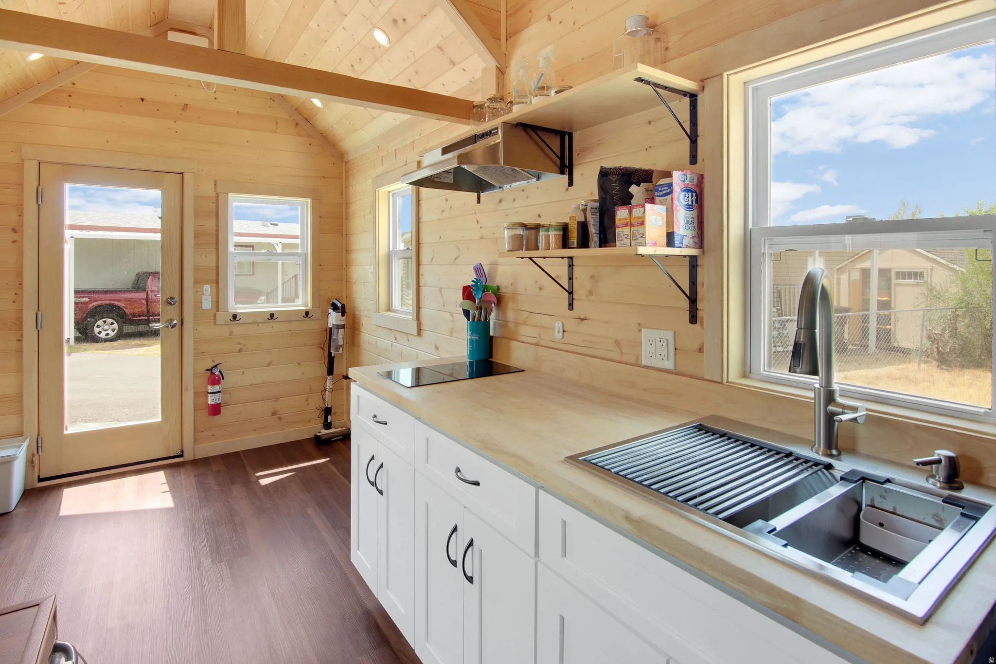 Kitchen featuring light wood-finish walls and vaulted ceiling, wood-finish flooring, white cabinetry with dark hardware, solid surface countertops, and a stainless steel sink with an integrated drying rack